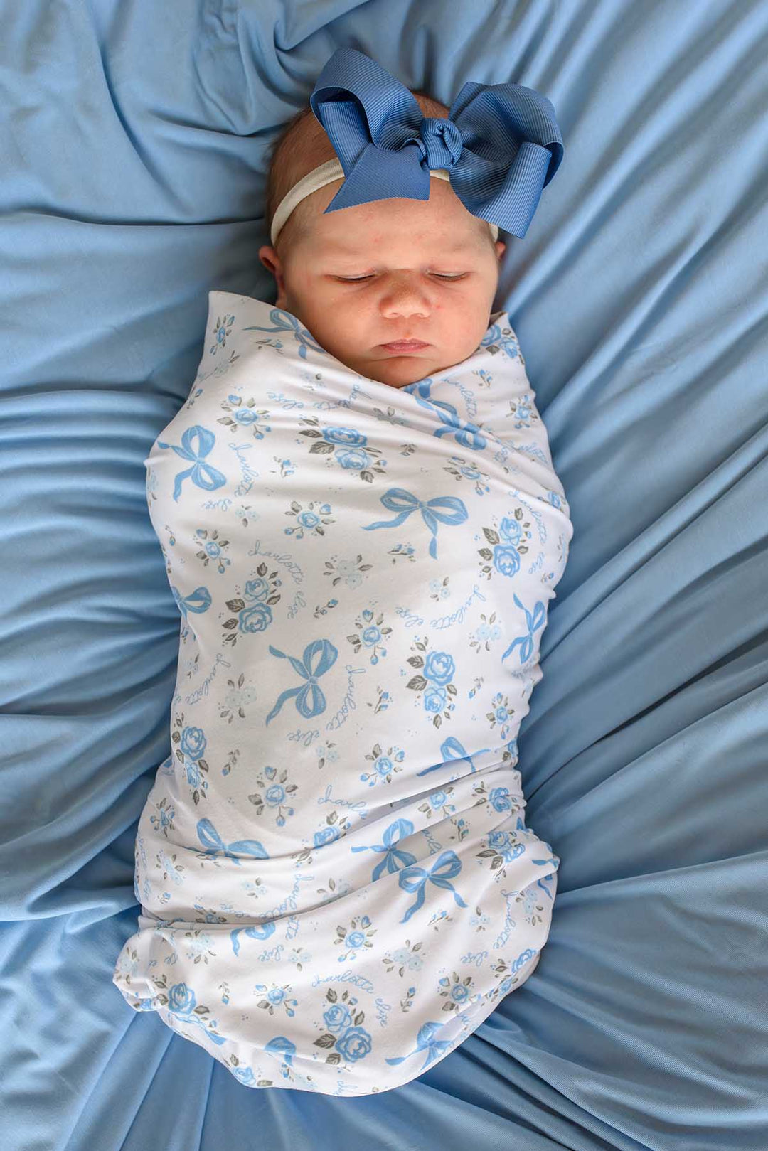 Overhead view of a newborn baby wearing a large blue bow, swaddled in a Tied In Blooms blanket with blue floral and bow print