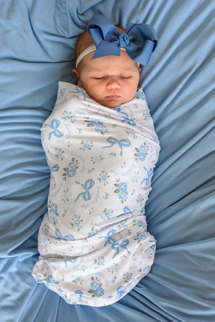 Overhead view of a newborn baby wearing a large blue bow, swaddled in a Tied In Blooms blanket with blue floral and bow print