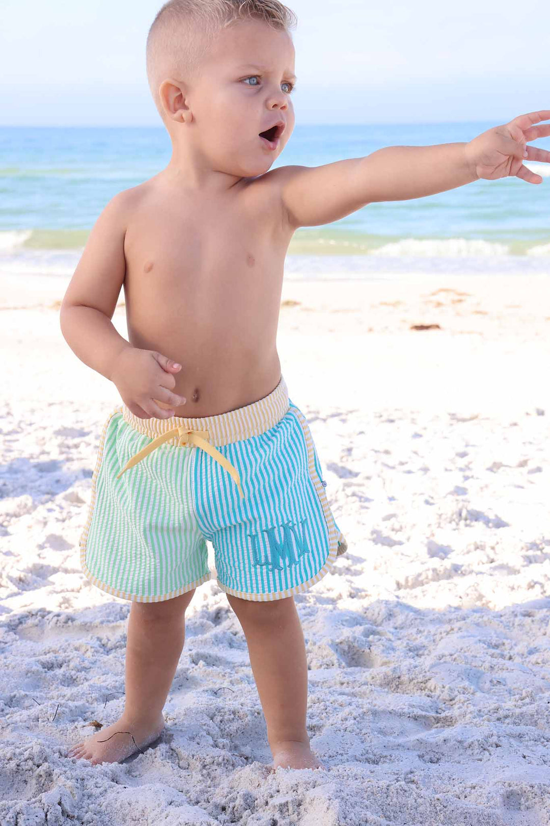 baby boy standing on beach in blue, green and yellow seersucker swim trunks 