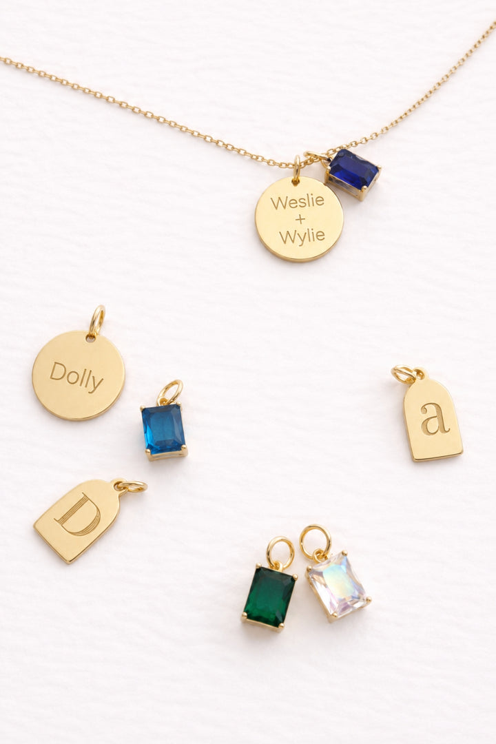 Caden Lane gold necklace with engraved "Weslie + Wylie" disc and blue birthstone charm, alongside "Dolly" name disc, initial "a" and "D" tag charms, and blue, green, and iridescent birthstone charms
