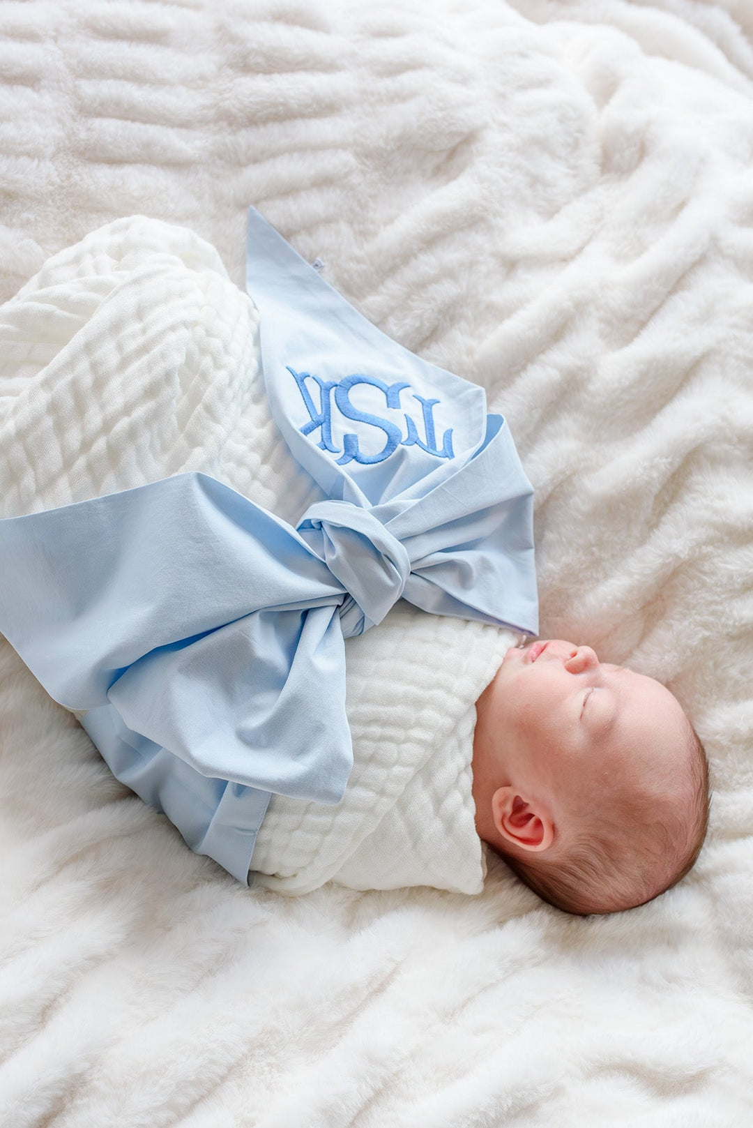 baby laying on fluffy white blanket wrapped in white muslin swaddle and light blue monogrammed bow sash