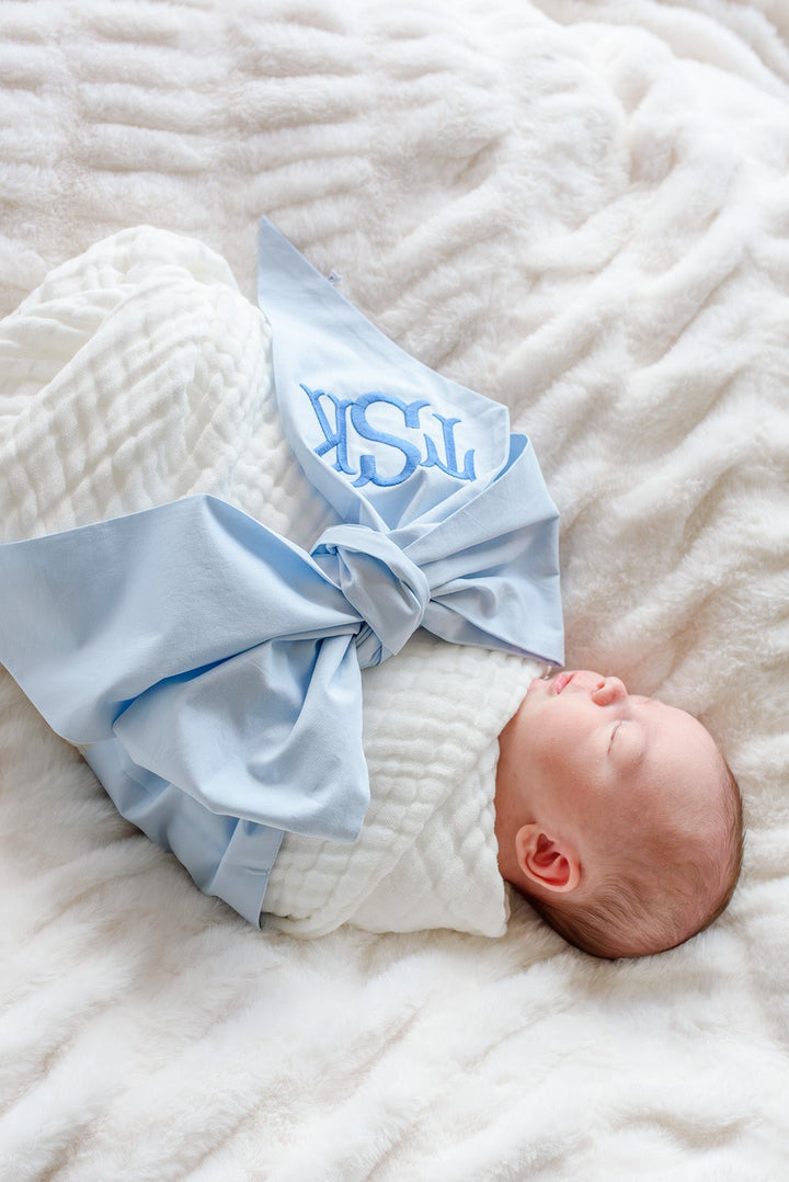 baby laying on fluffy white blanket wrapped in white muslin swaddle and light blue monogrammed bow sash