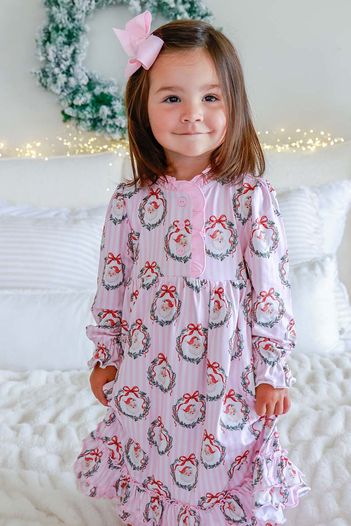 Girl in a Caden Lane pink striped nightgown with vintage Santa wreath print, sitting on a bed with a pink bow in her hair