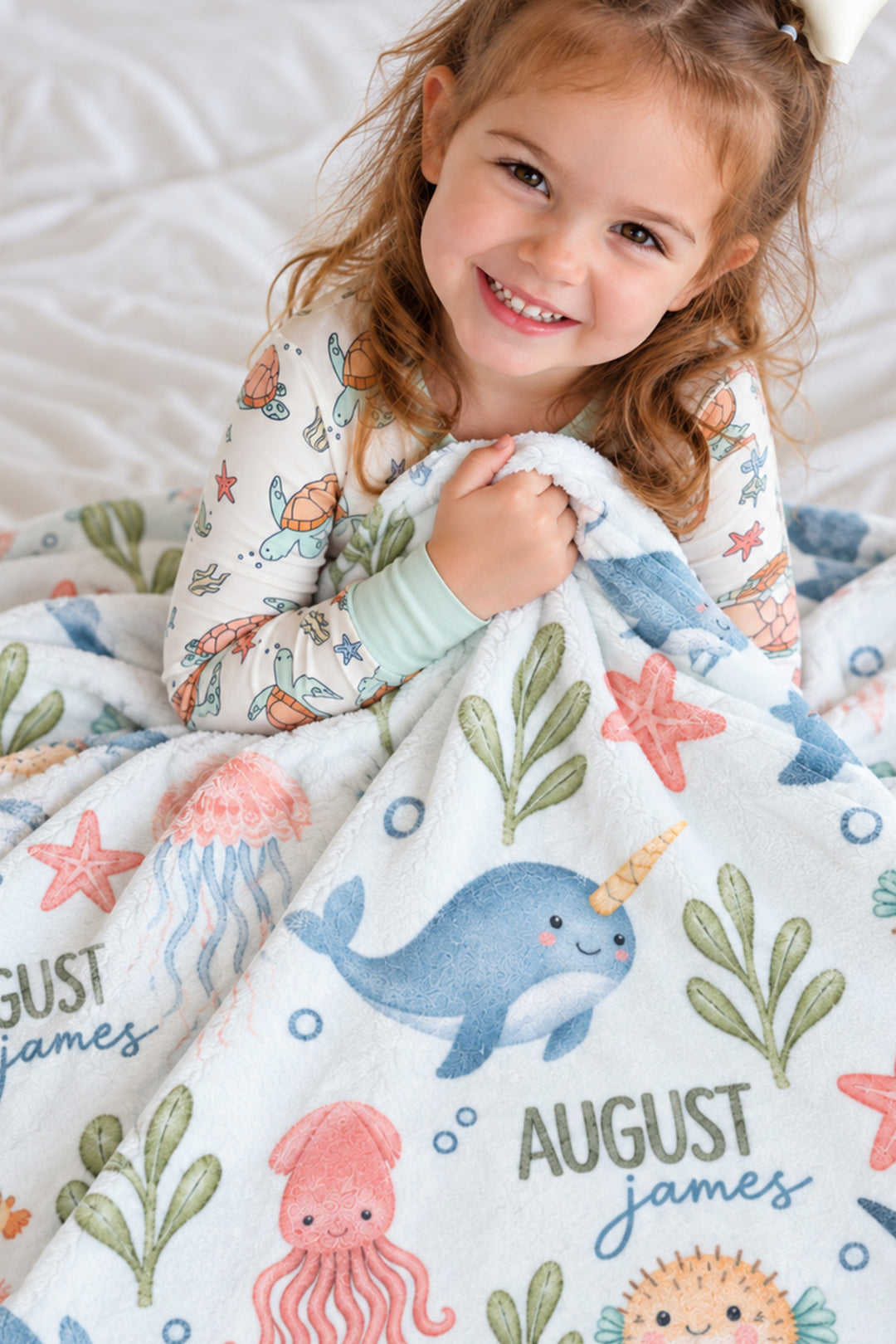Smiling girl in sea turtle-print pajamas holding up a personalized Caden Lane Ocean Dreams blanket, showing narwhal, jellyfish, and custom name print