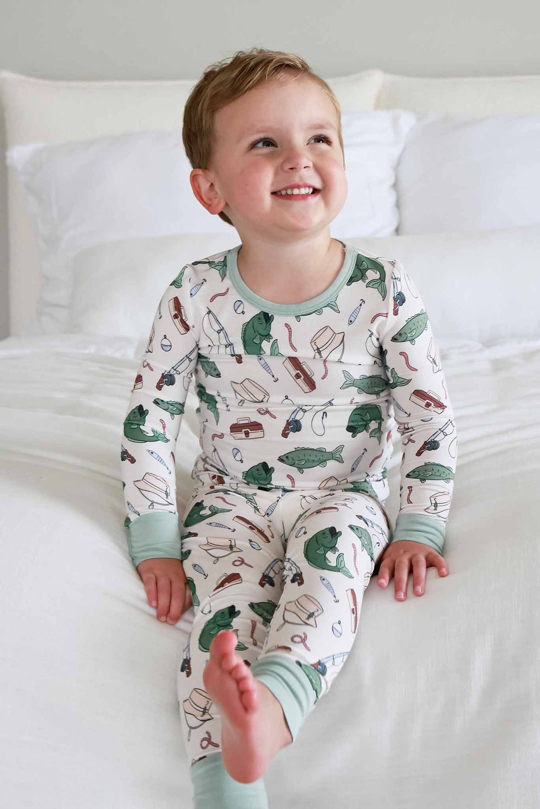 Smiling toddler on bed wearing the Off The Hook two piece pajama set showing full length with fishing print