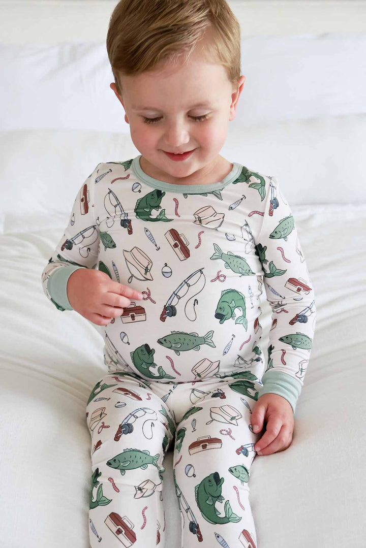 Toddler sitting and looking down wearing the Off The Hook two piece pajama set showing fishing print detail