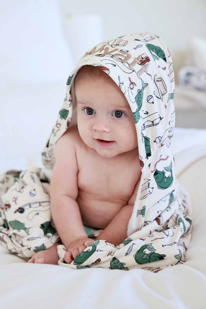 Baby sitting up wearing the Off The Hook embroidered bamboo hooded towel with fishing themed print
