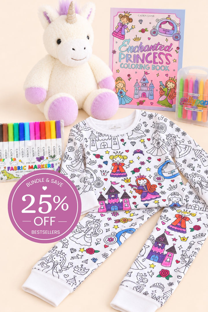 Flat lay of a Caden Lane color-in princess pajama set with colored castles and princesses, paired with a plush unicorn, fabric markers, crayons, and a princess coloring book.