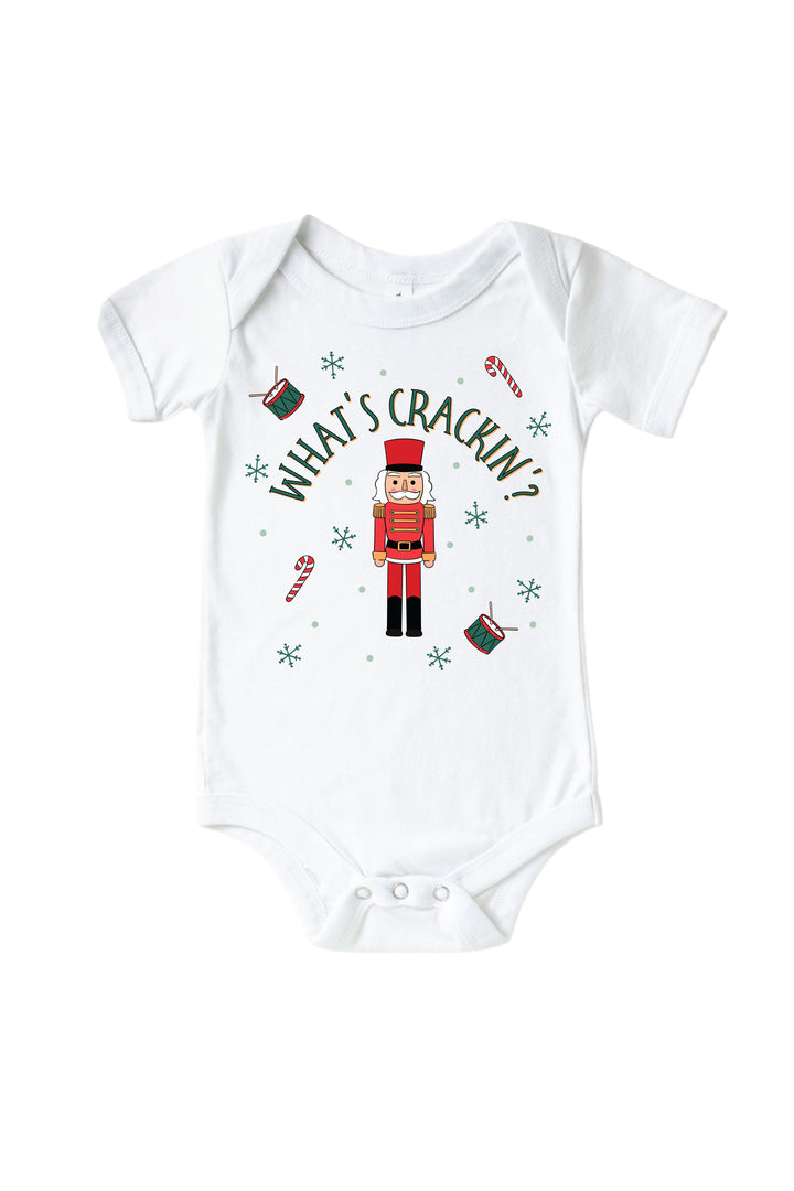 Caden Lane's white "What's Crackin'" baby onesie featuring a red and green toy soldier nutcracker with snowflakes, candy canes, and drums, laid flat.
