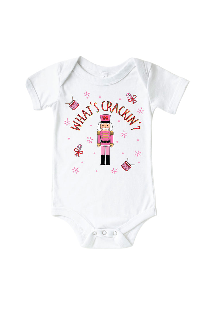 Caden Lane's white "What's Crackin'" baby onesie featuring a pink toy soldier nutcracker with snowflakes, candy canes, and drums, laid flat.