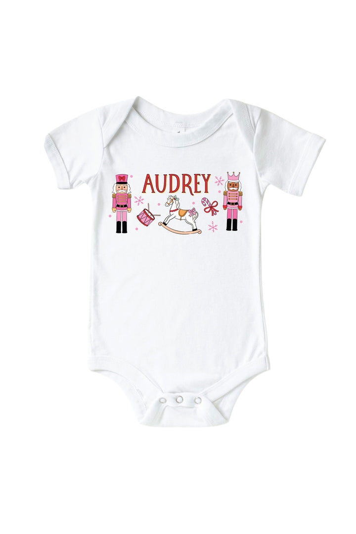 Caden Lane's white baby onesie personalized with the name "Audrey," featuring pink toy soldier nutcrackers, a rocking horse, drum, and candy cane, laid flat.