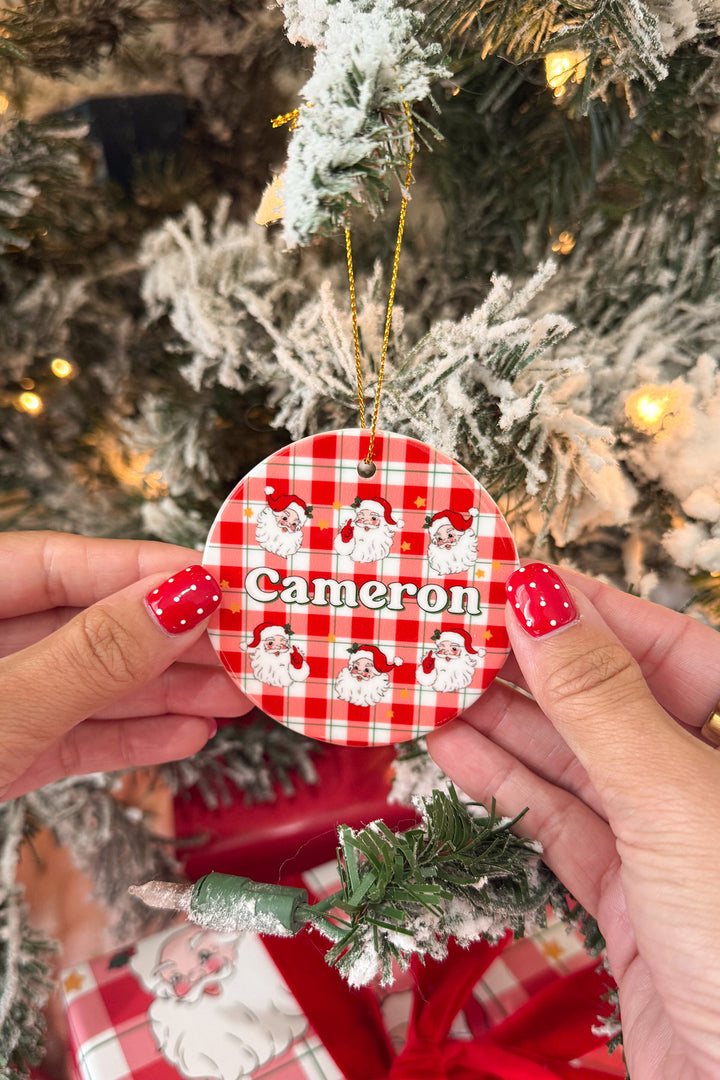 A round Christmas ornament held by two hands in front of a decorated tree. The ornament has a red and white gingham pattern with Santa illustrations and the name 'Cameron' in white script.