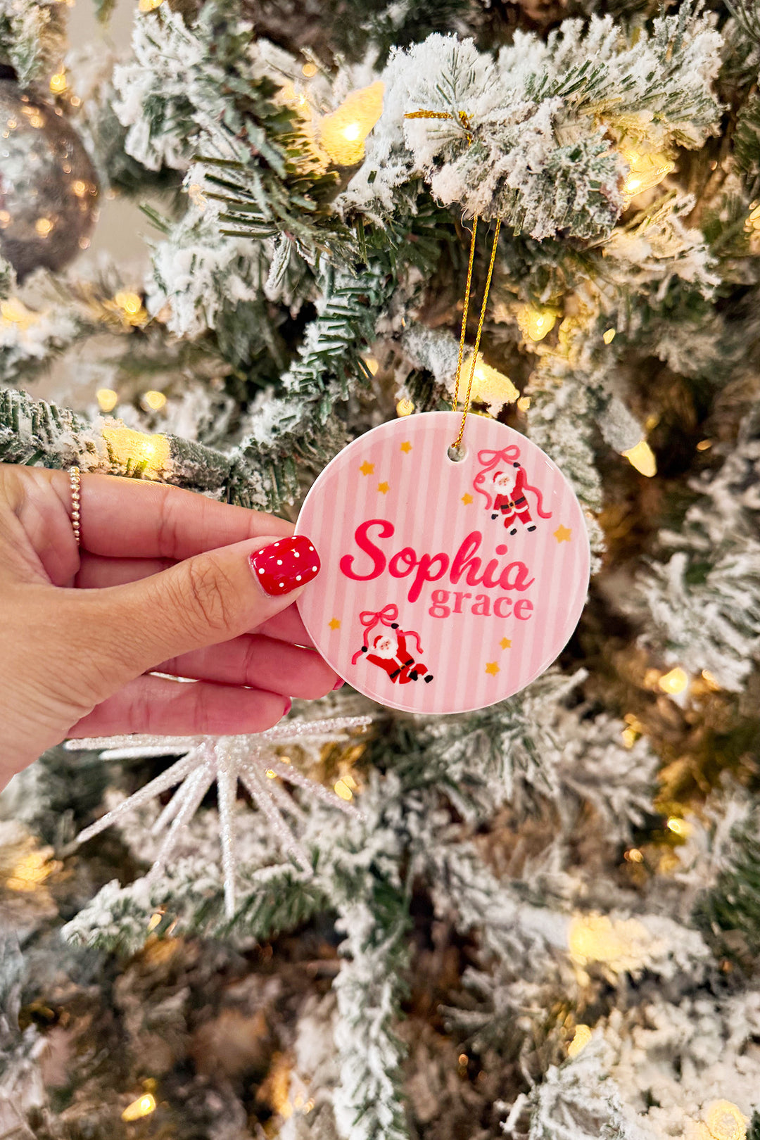 A hand  holds a round pink striped Caden Lane Christmas ornament hanging from a flocked Christmas tree with warm lights. The ornament is personalized with the name 'Sophia grace' in red script and small red and black illustrations.