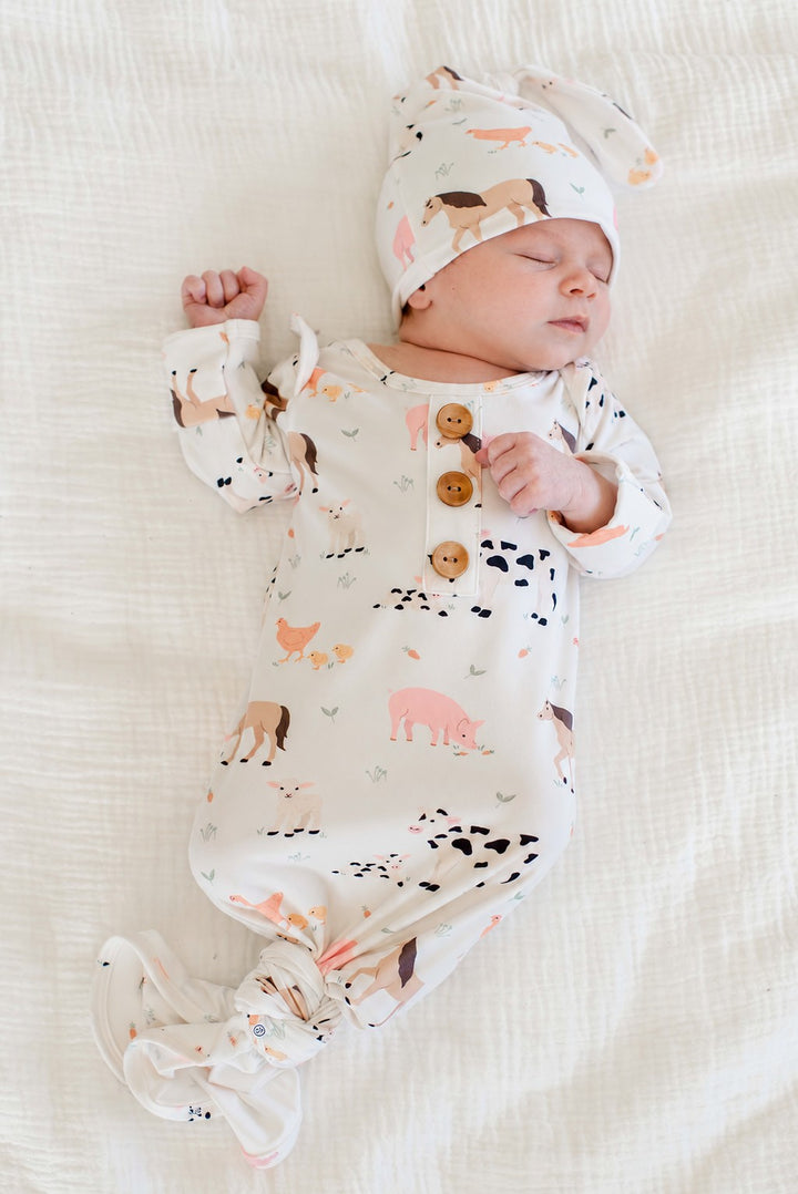 Sleeping newborn wearing Caden Lane farm animal knotted gown with wooden buttons and matching topknot hat, lying on white bedding