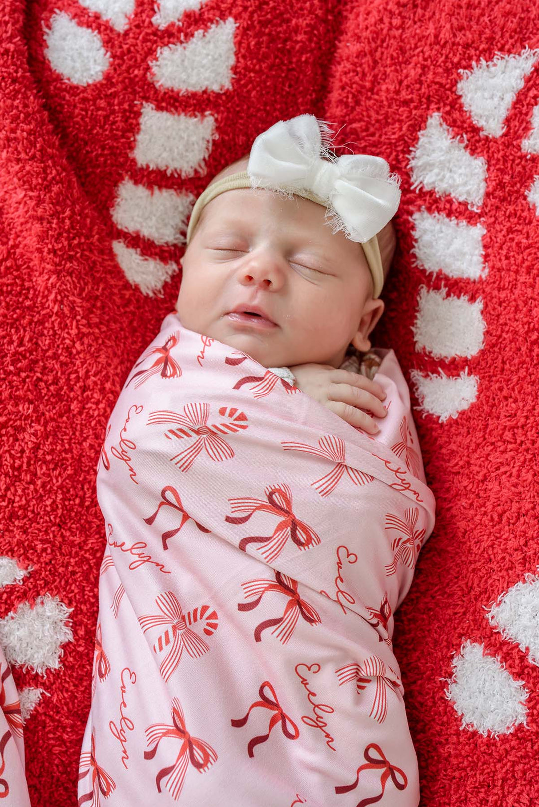 Newborn wrapped in the Caden Lane Personalized Peppermint Bows Christmas Swaddle Blanket featuring festive red bows, candy canes, and a custom name on a soft blush pink background.