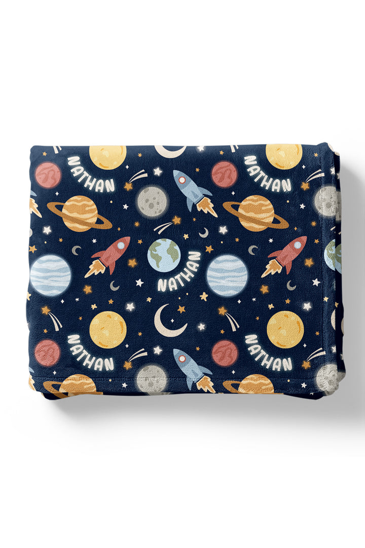 Flat lay of a neatly folded Caden Lane navy plush blanket in a Stellar Snuggles print featuring Saturn, Earth, rockets, and crescent moons, reading “Nathan.”
