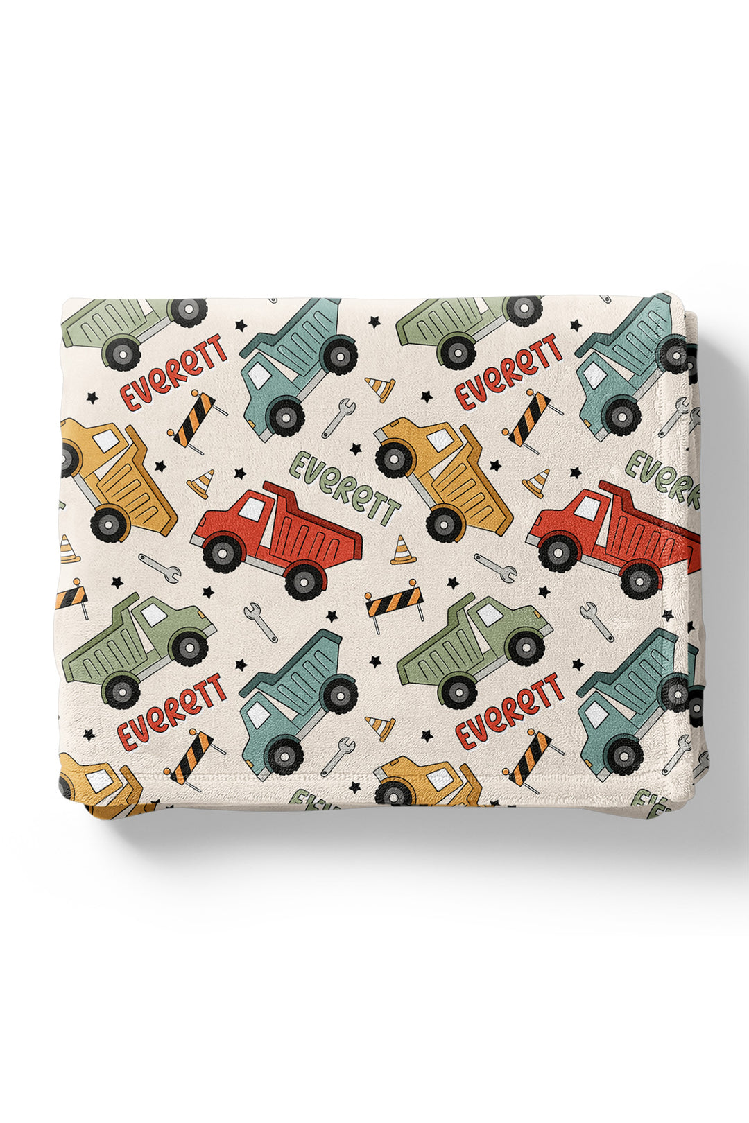 Flat lay of a folded Caden Lane cream plush blanket in a Busy Builder print featuring tilted dump trucks, cones, and barricades, reading "Everett" in red and green.