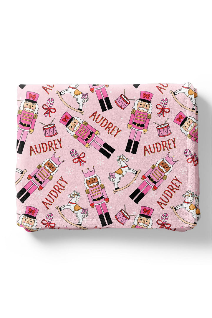 Personalized Caden Lane Christmas blanket featuring pink nutcrackers, rocking horses, drums, bows, snowflakes, and a custom child’s name.