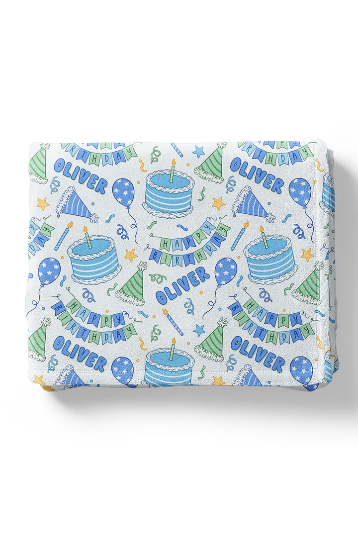 A Caden Lane personalized kids blanket featuring a white background with a colorful 'Happy Birthday Oliver' party pattern, including blue and green illustrations of cakes, balloons, hats, and banners.
