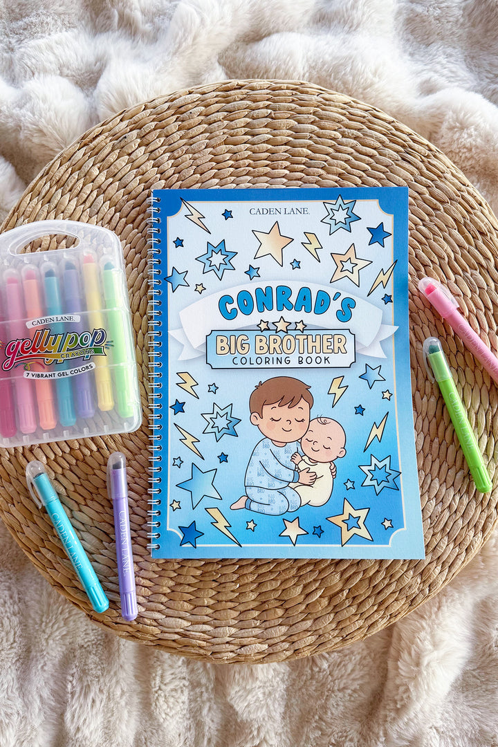 Caden Lane 'Conrad's Big Brother Coloring Book' on a woven tray with a cream throw, Gellypop gel crayon case, and pastel Caden Lane markers.