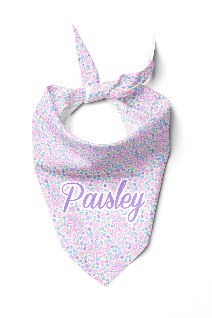 Personalized pet bandana in the Ribbon Garden print — a soft pink and lavender floral pattern — tied in a knot and displayed flat, personalized with the name "Paisley"