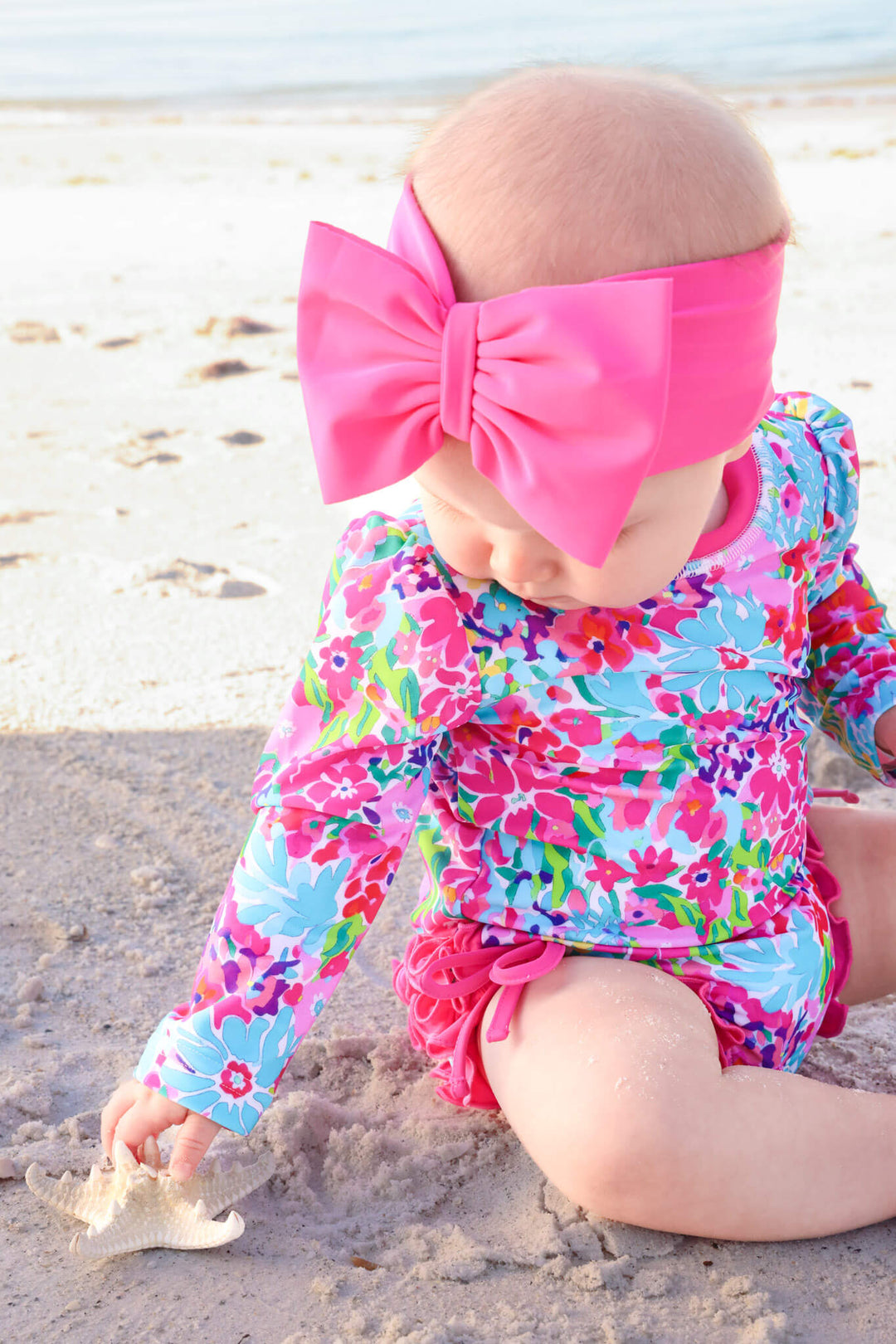 baby girl sitting on beach in petal splash long sleeve ruffle bottom rash guard with matching pink swim bow