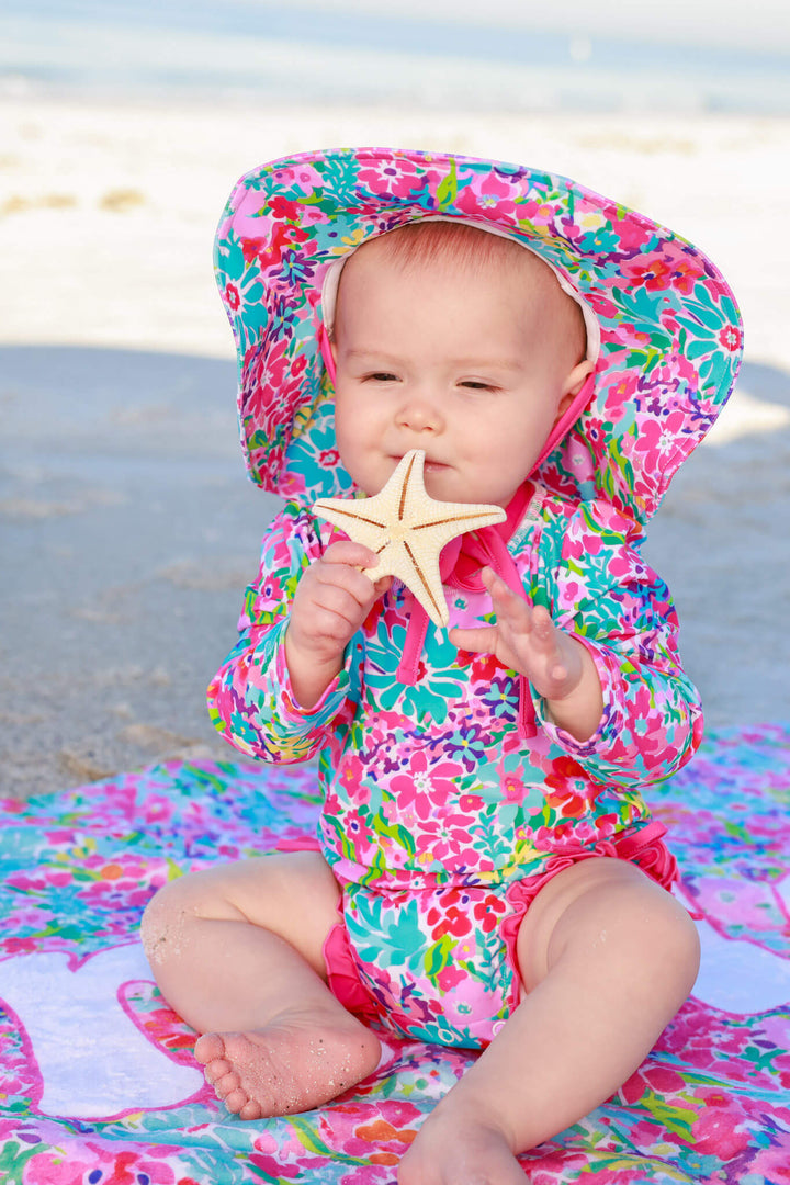 baby girl sitting on beach playing with starfish wearing petal splash sun hat, swimsuit and towel