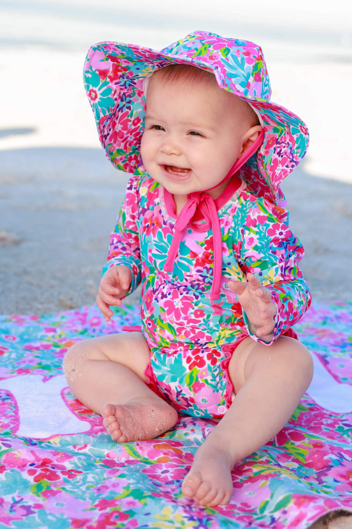 baby girl sitting on beach in petals splash ruffle bottom rash guard, sun hat and personalized towel 