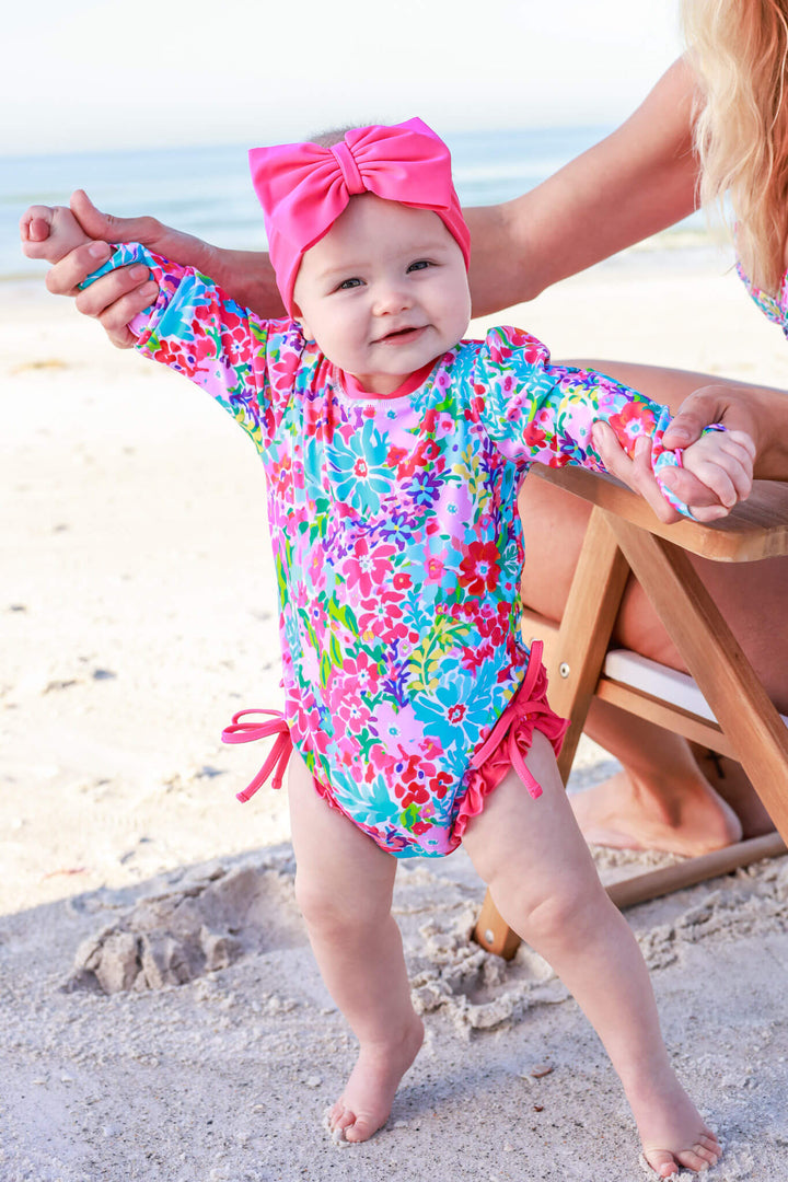 baby girl walking on the beach in long sleeve rash guard with ruffle bottom in petal splash print with a matching pink swim bow