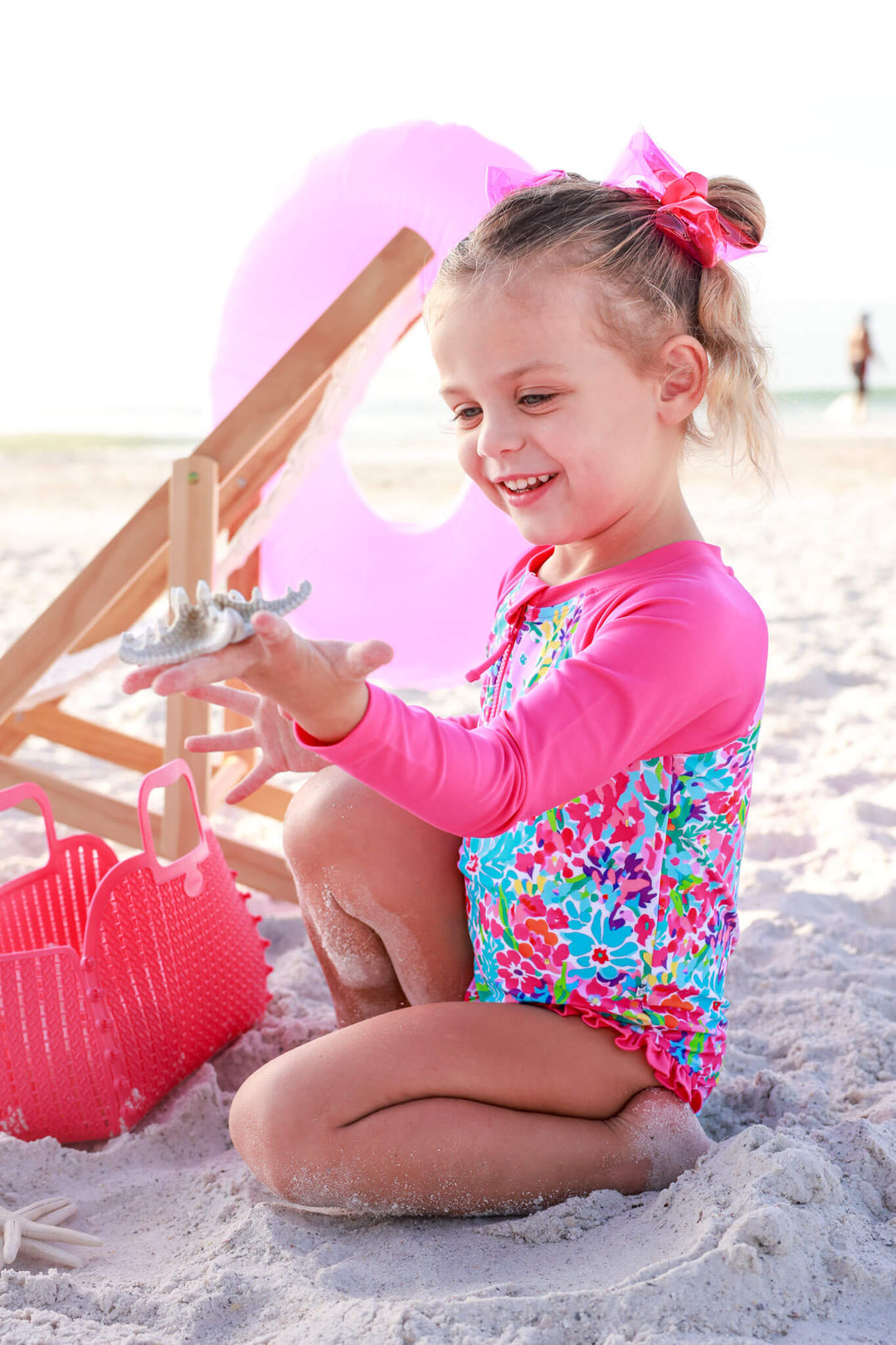 girl playing on the beach in petal splash long sleeve rash guard swimsuit with front zipper with pink ribbon in her hair 