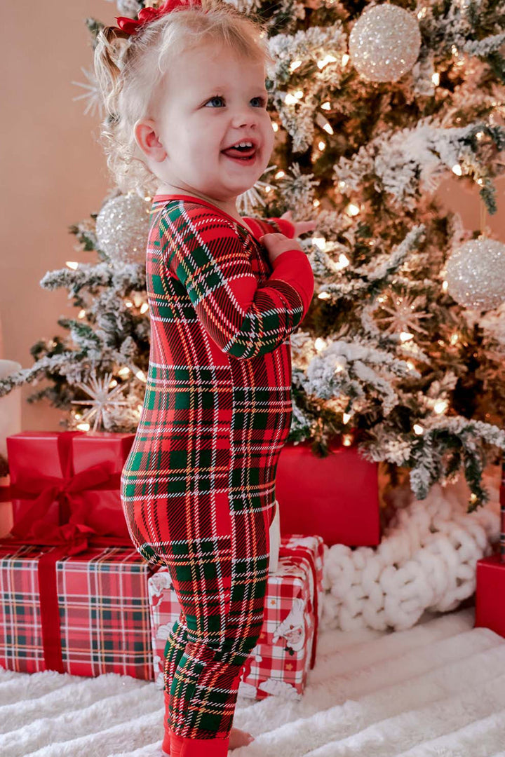 Caden Lane toddler wearing a red and green plaid bamboo Christmas zipper pajama romper, standing beside a decorated Christmas tree and wrapped holiday gifts in matching family pajamas.