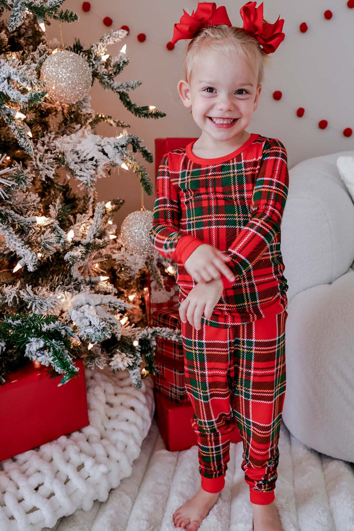 Toddler wearing Caden Lane Christmas bamboo plaid pajamas in a festive red and green tartan print, standing by a decorated Christmas tree in matching holiday sleepwear.