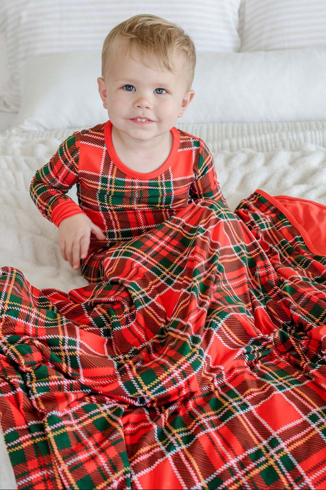 Caden Lane toddler wearing red and green plaid bamboo Christmas pajamas cuddled in a matching plaid blanket, creating a cozy holiday scene perfect for Christmas morning, family photos, and festive traditions.