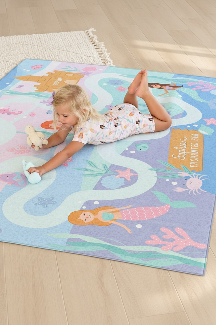 Young girl playing on a personalized Caden Lane mermaid playroom rug featuring pastel mermaids, sea creatures, coral and an underwater castle.