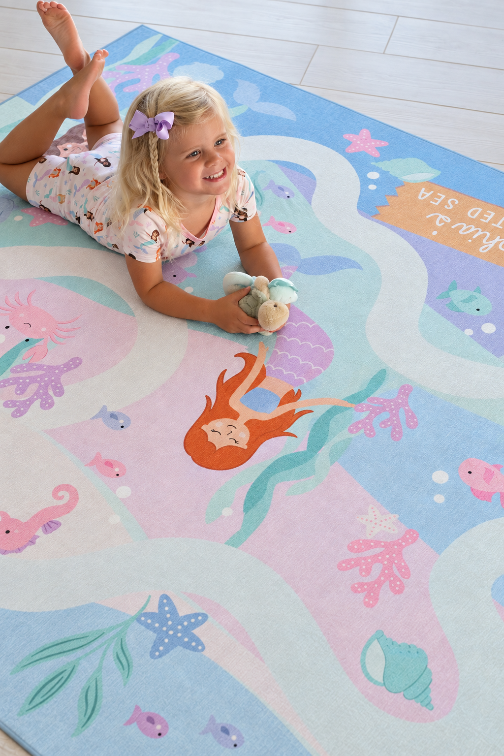 Young girl sitting on a Caden Lane mermaid playroom rug decorated with pastel mermaids, fish, starfish, coral and winding ocean paths.