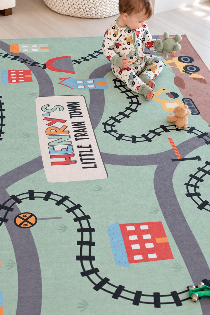 Baby sitting on a personalized Caden Lane train-themed playroom rug with colorful tracks, roads, buildings and construction vehicles.