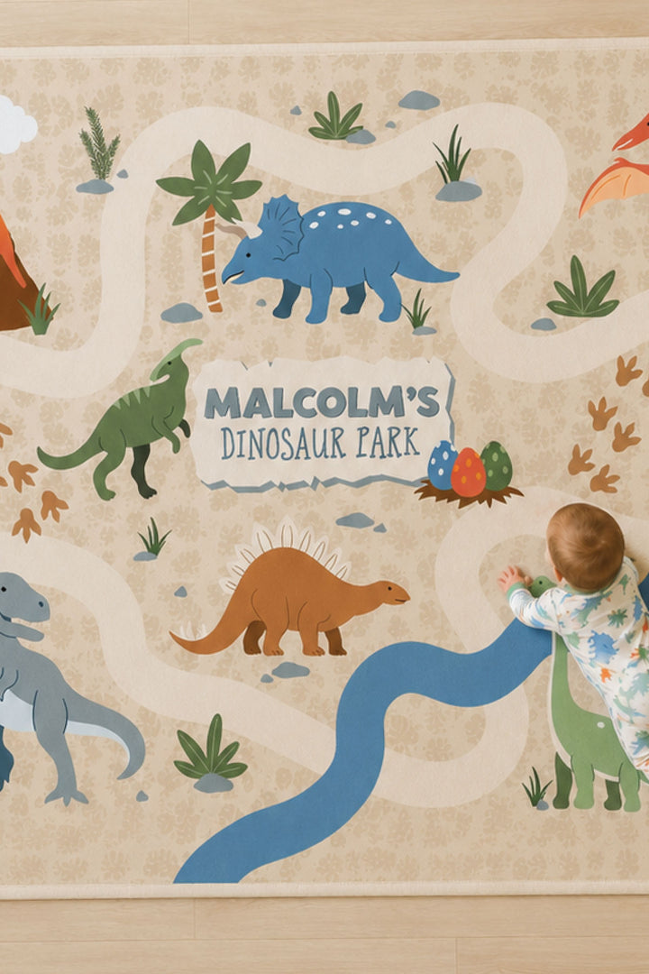Close-up of a Caden Lane personalized dinosaur play rug showing a blue triceratops, brown stegosaurus, dinosaur eggs, and a winding path around the text 'Malcolm's Dinosaur Park,' with a baby crawling on the rug in a dinosaur-print onesie