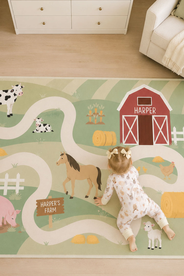 Close-up of a Caden Lane personalized farm play rug showing a red barn labeled 'Harper,' a horse, cows, chickens, and a wooden sign reading 'Harper's Farm,' with a baby in bows and a floral onesie crawling across it