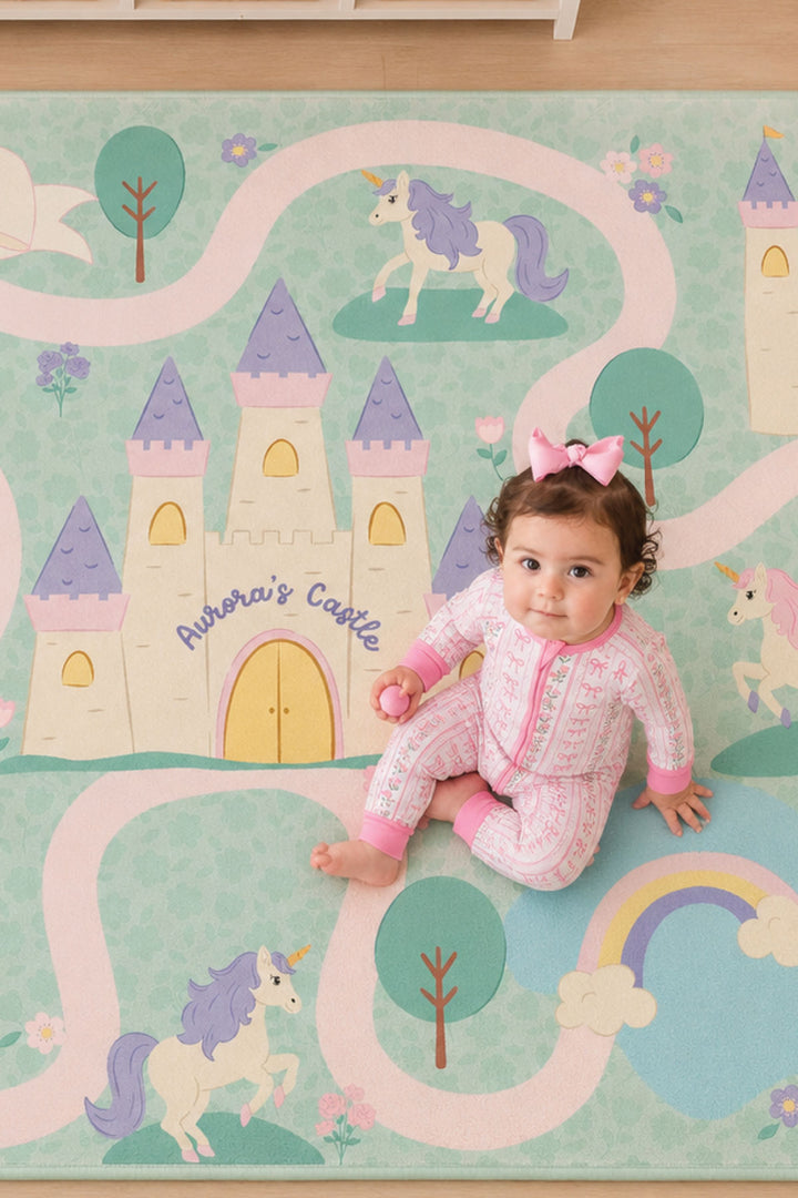 Close-up of a Caden Lane personalized princess play rug showing a pastel castle labeled 'Aurora's Castle,' unicorns, trees, and a rainbow, with a baby wearing a pink bow and bow-print pajamas sitting on the rug