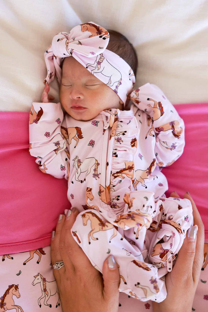 newborn girl laying on bamboo blanket in girly pony ruffle zipper footie with matching headwrap