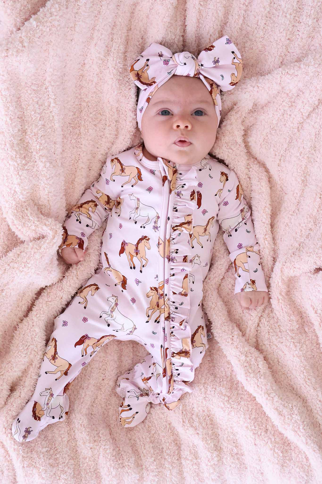 baby girl laying on fluffy pink blanket wearing the pony club pink ruffle zipper footie with matching headwrap 
