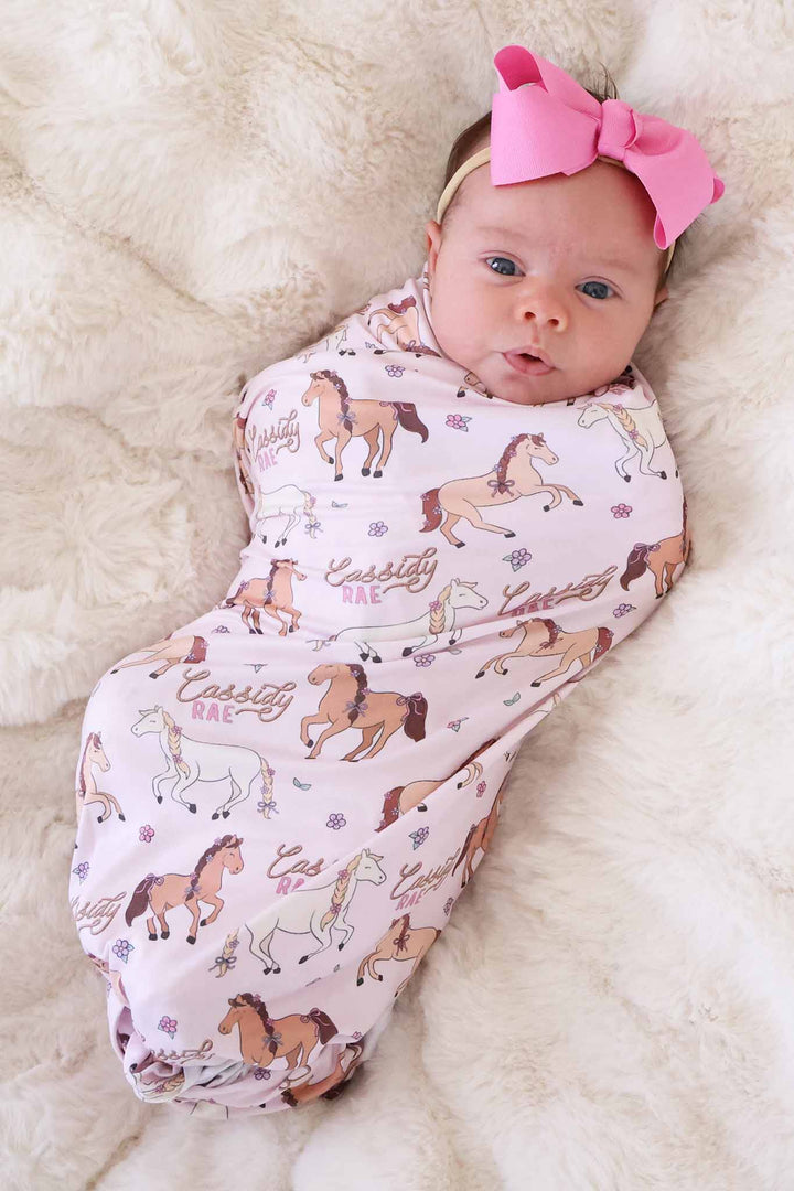 newborn girl wrapped in pink pony club personalized swaddle blanket with matching pink ribbon bow headband 