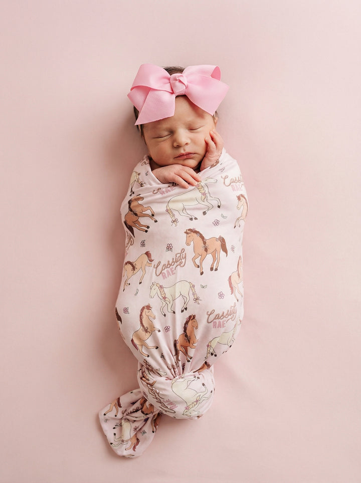 baby girl wrapped in pony club pink personalized swaddle blanket with matching pink ribbon bow headband on a pink background 