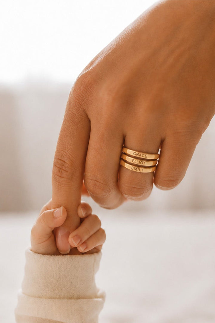 Caden Lane personalized gold name rings engraved with Grace, Elliot, and Emily stacked on a mother's finger while holding a baby's hand.