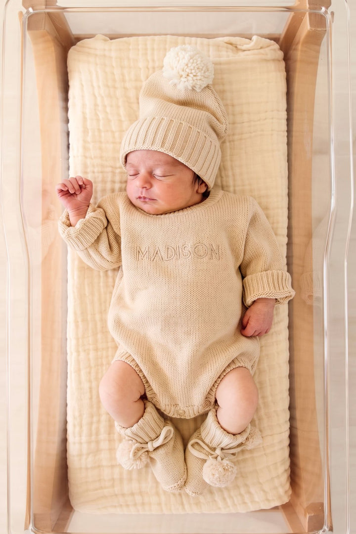 A sleeping newborn baby wearing a beige knit hat with a pom-pom, a matching long-sleeve romper with the name 'MADISON' embroidered on the chest, and matching booties, lying on a textured cream muslin blanket inside a clear baby bassinet.