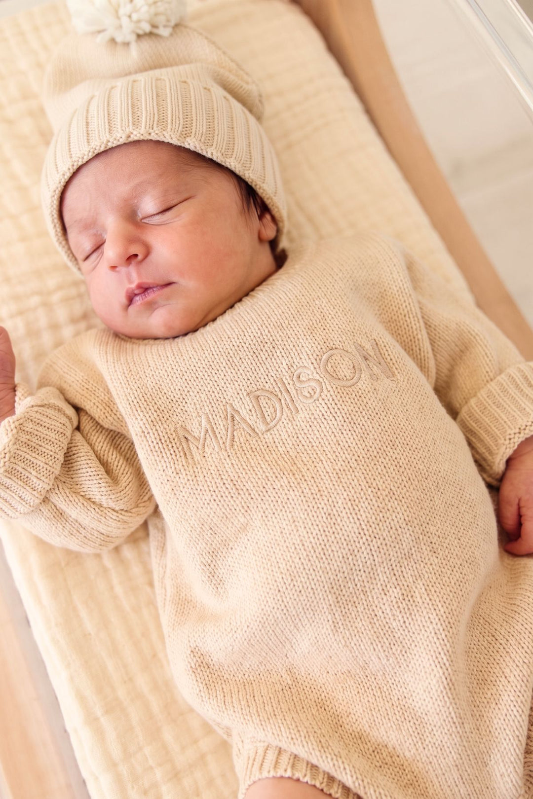 A sleeping newborn baby wearing a beige knit beanie with a pom-pom and a matching long-sleeve knit romper with the name 'MADISON' embroidered on the chest, lying on a textured cream muslin blanket.