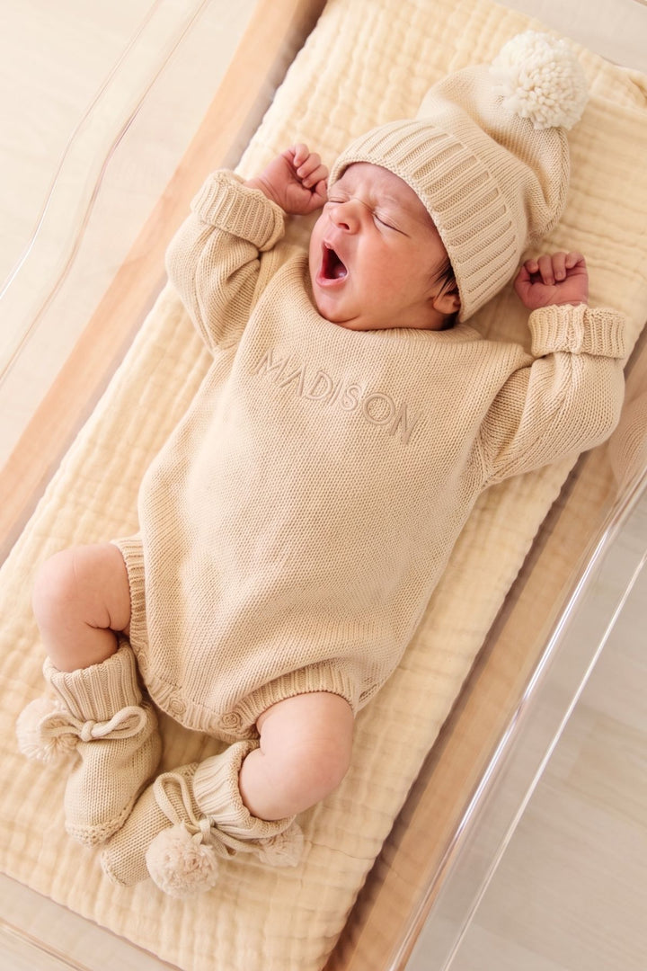 Yawning newborn in a bassinet wearing Caden Lane sand-colored personalized knit romper "Madison," pom-pom hat, and matching pom-pom tie booties