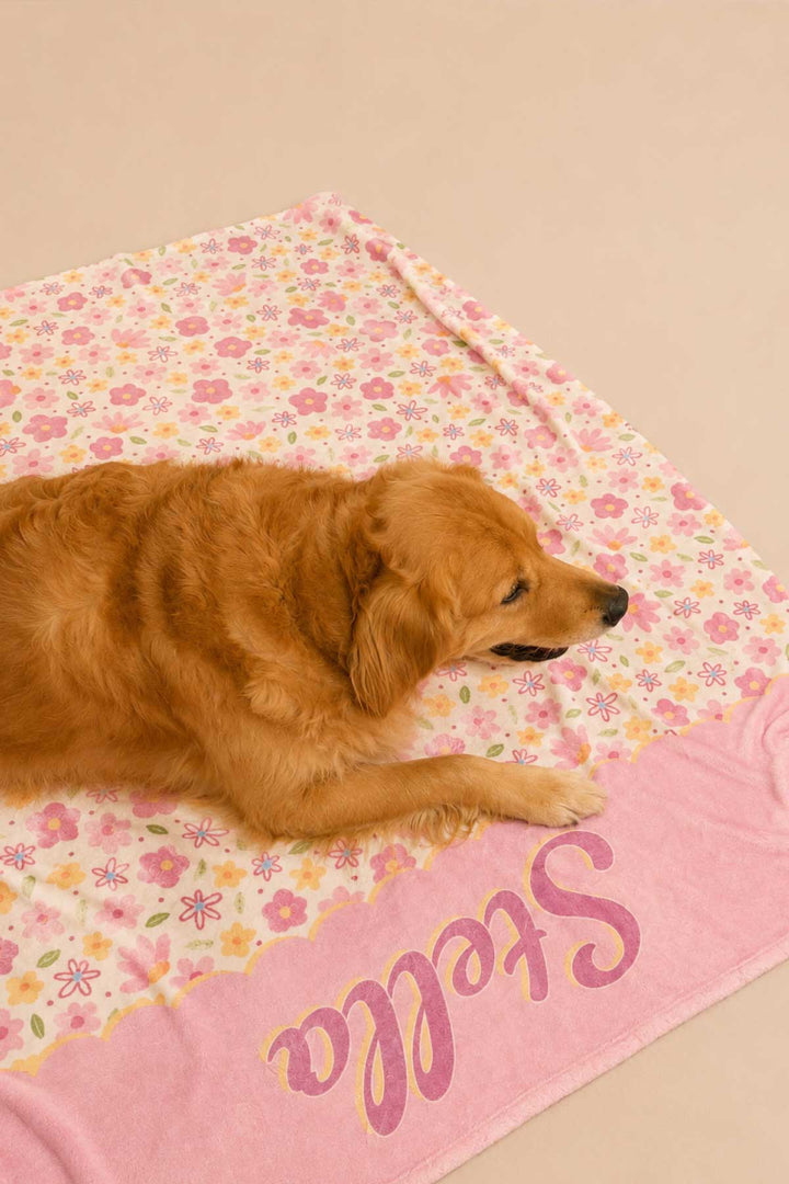 Golden retriever lying on a Scattered Blooms personalized pet blanket, showing the colorful pink, purple, and yellow floral print with pink personalized name panel