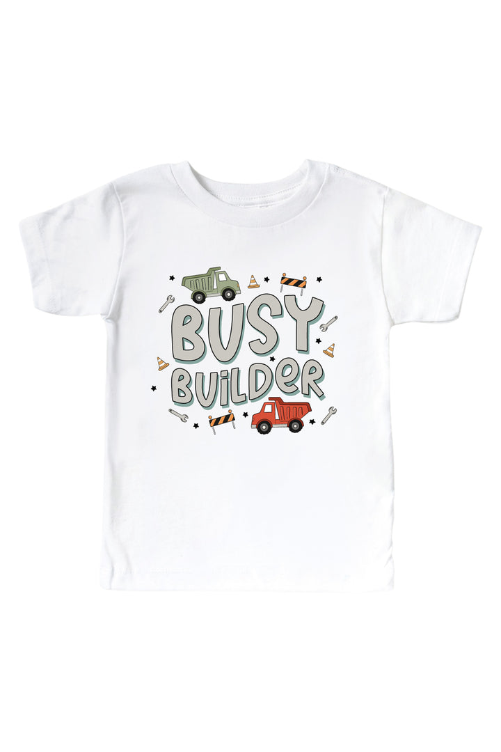 Flat lay of a Caden Lane white short-sleeve tee in a Busy Builder print featuring dump trucks, wrenches, and barricades scattered around the words "Busy builder."