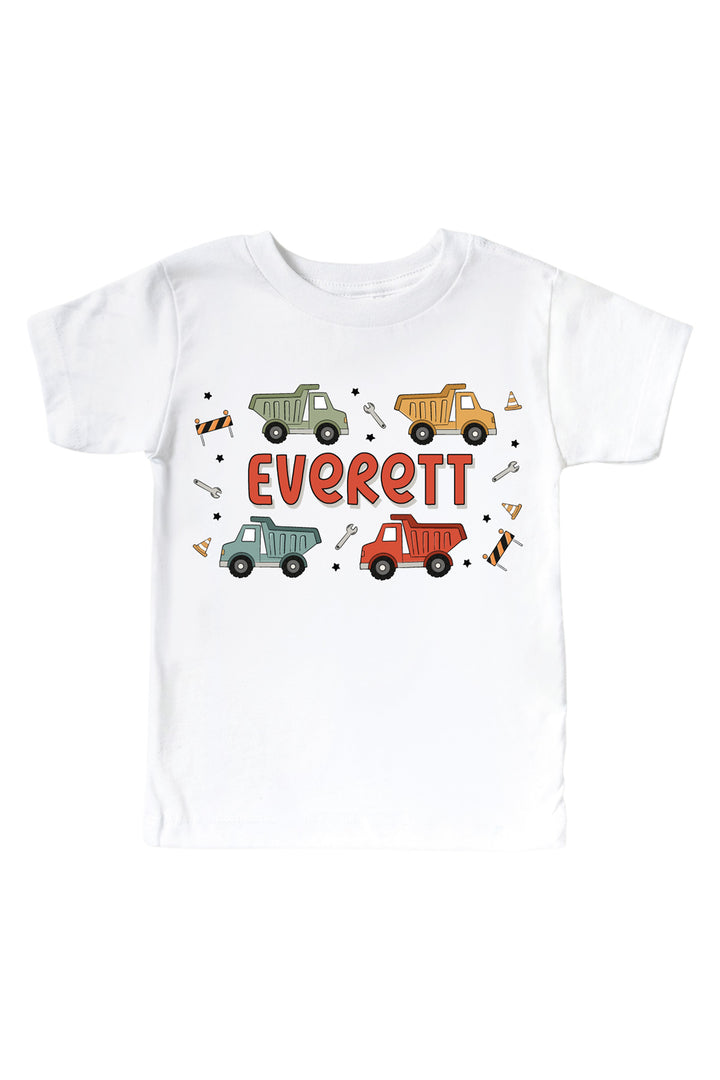 Flat lay of a Caden Lane white short-sleeve tee in a Busy Builder print featuring four dump trucks, wrenches, and traffic cones, reading "Everett" in red.